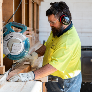 Worker in hi-vis and safety gear cutting timber with a mitre saw in a workshop.